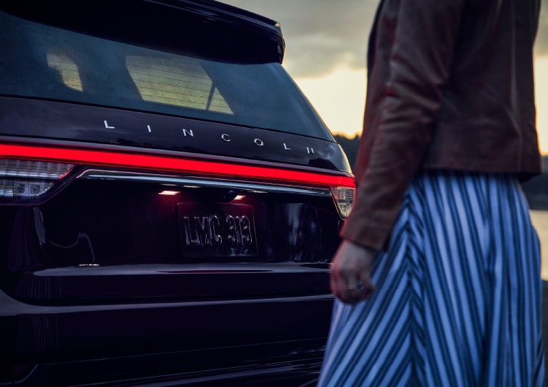 A person is shown near the rear of a 2024 Lincoln Aviator® SUV as the Lincoln Embrace illuminates the rear lights | Rogers Lincoln in Midland TX